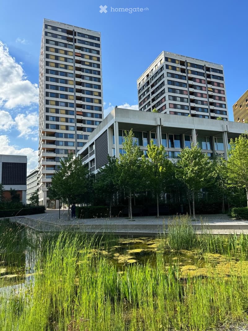 Modern residential complex with two tall apartment buildings, surrounded by greenery and a small water feature in the foreground.