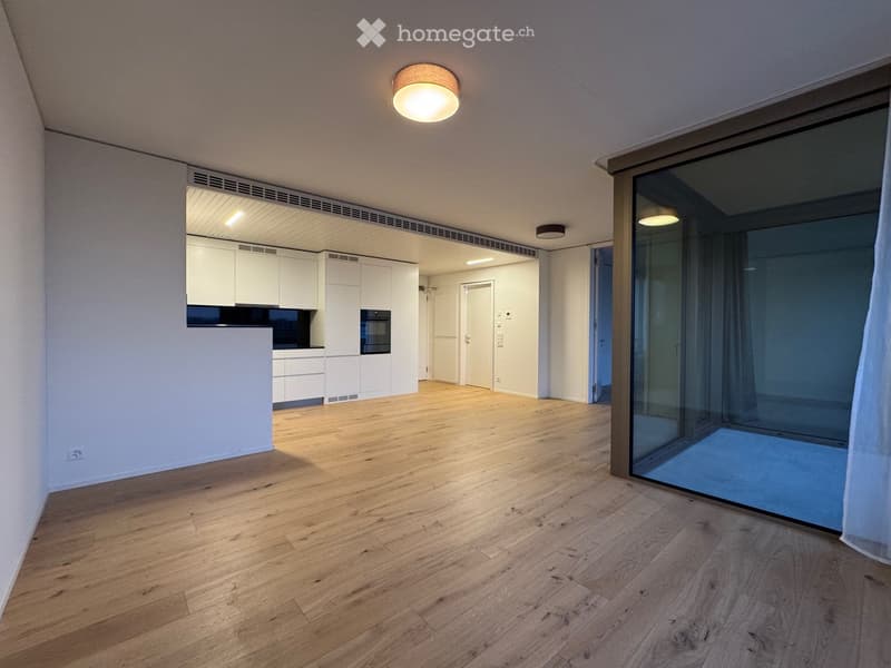 Open-plan unfurnished room with wooden flooring, white walls, and a large glass door leading to a balcony. A small kitchen area is visible in the background with white cabinets and a built-in oven.