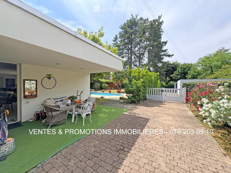 Outdoor patio area with wicker chairs, a small table, artificial grass, brick pathway, and a swimming pool in the background.
