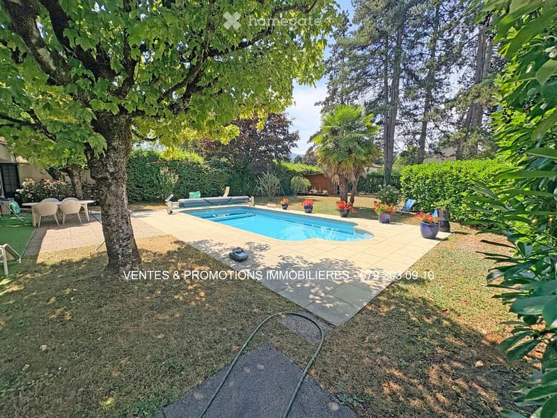 A well-maintained garden with a swimming pool, surrounded by trees, shrubs, and potted plants. There are outdoor chairs and a table near the pool area.