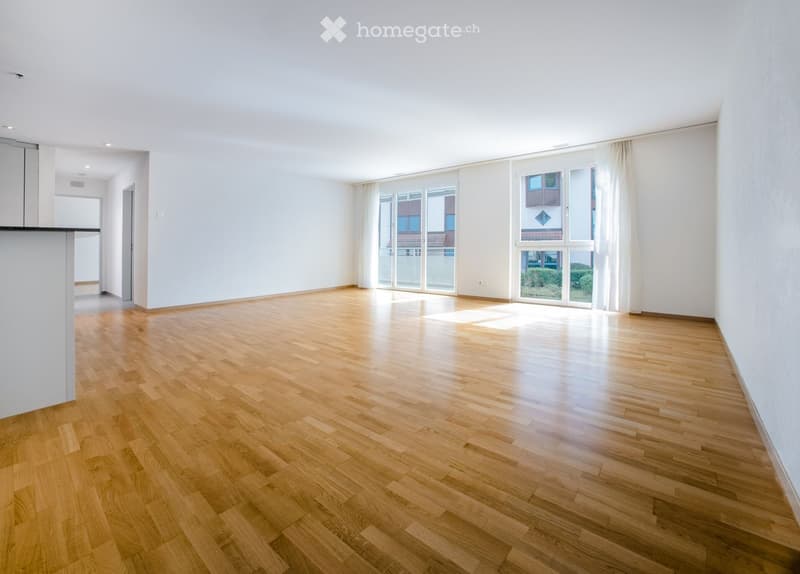 Empty room with wooden flooring, white walls, large windows with curtains, and a doorway leading to another area.