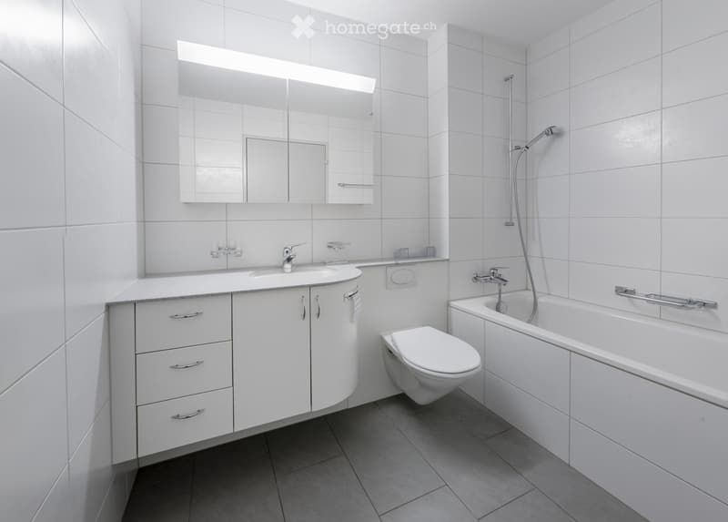 Modern bathroom with white tiles, a sink with a mirror, a bathtub, and a toilet. The space features sleek cabinetry and a modern faucet.
