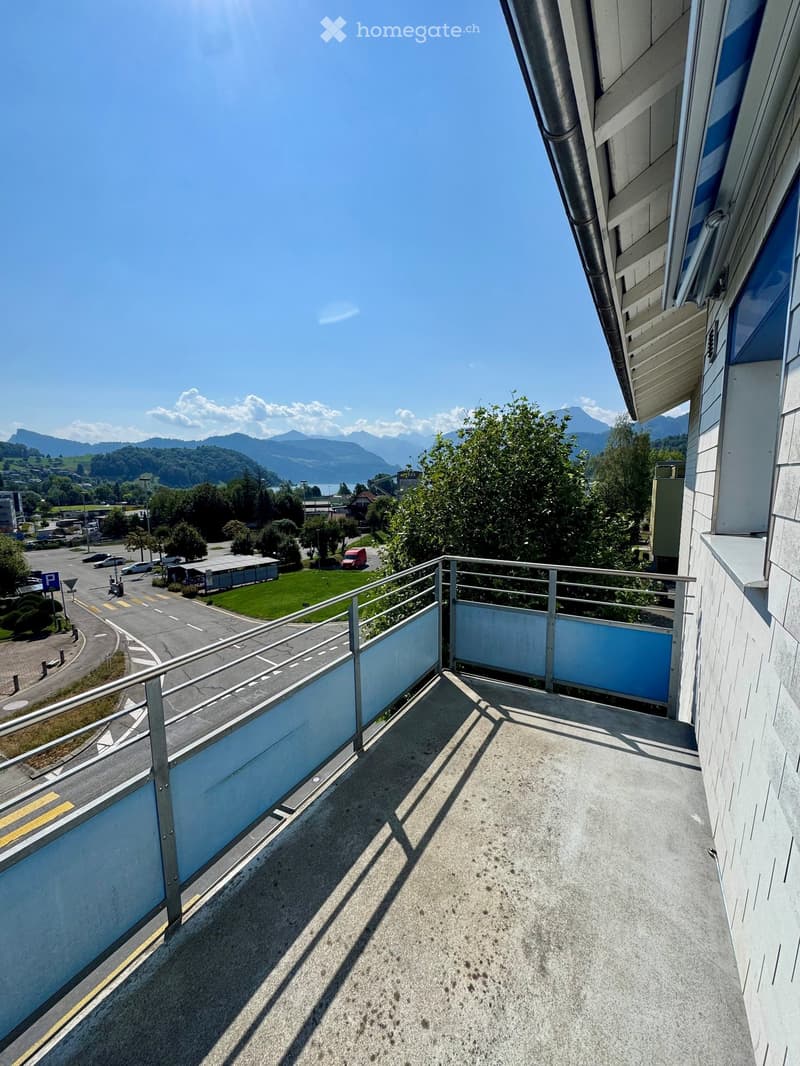 Balcony with metal railing, overlooking a road, parking area, and distant mountains.