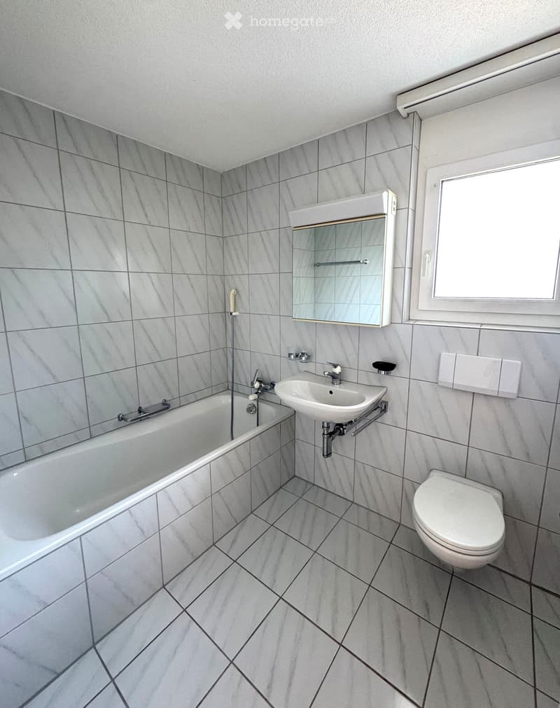 Modern bathroom with grey tiled walls and floor, featuring a bathtub, sink, toilet, and a mirror above the sink.
