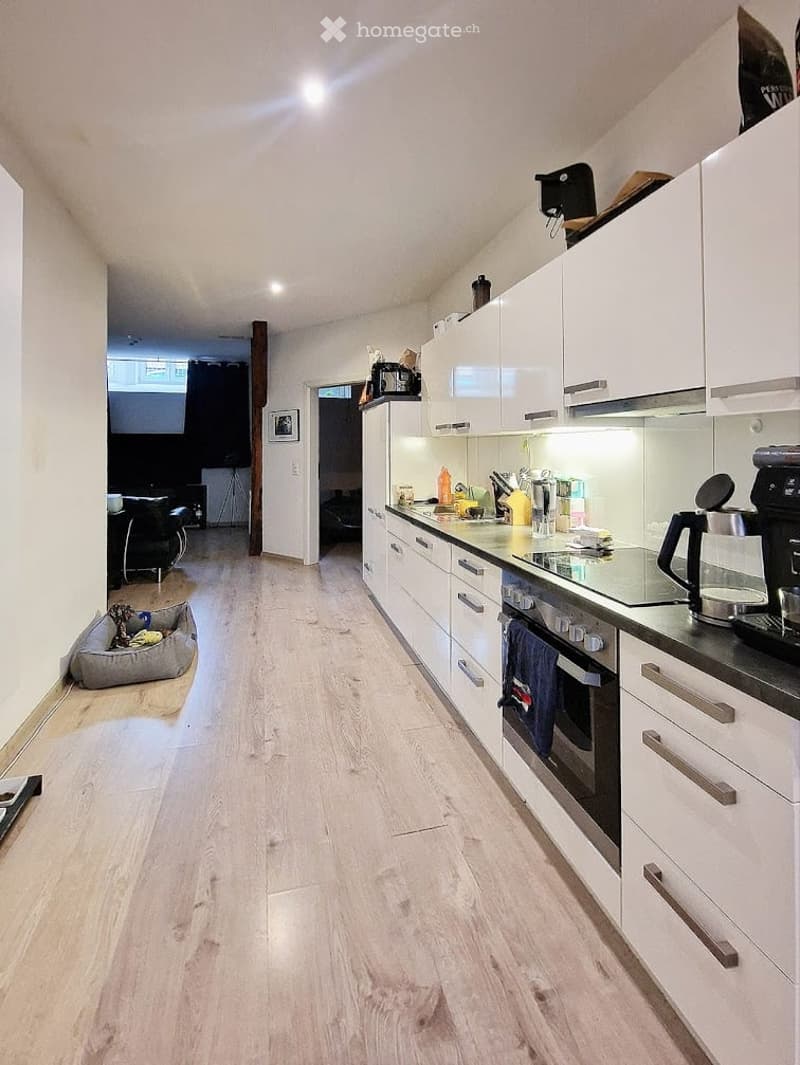 Modern kitchen with white cabinets, stainless steel appliances, and a light wooden floor. Visible items include an oven, coffee maker, microwave, and a small kitchen island.