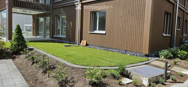 Garden / Outdoor Area. Well-maintained garden with a neatly trimmed lawn, small trees, shrubs, and a paved pathway leading to a modern building with large windows and a balcony.