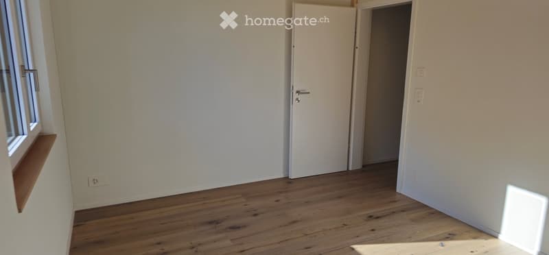 Empty room with wooden flooring, white walls, a window with a wooden sill, and a closed white door leading to another space.