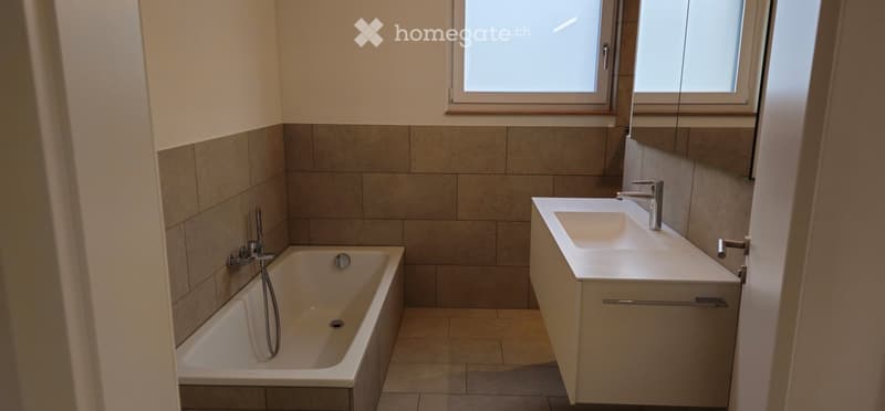 Modern bathroom with a bathtub, sink, and large mirror. The walls are tiled in neutral tones, and the floor is tiled as well. There are two windows providing natural light.