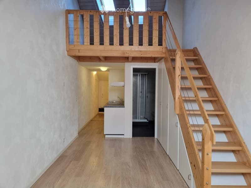 A narrow hallway with wooden flooring, leading to a small kitchen area and a staircase to an upper loft space. The loft has a wooden railing and skylight windows.