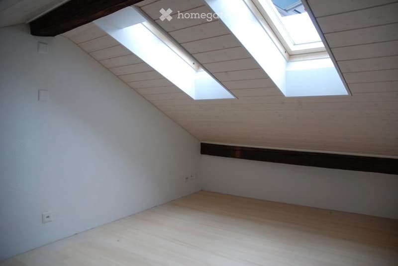 Empty attic room with wooden floor, white walls, skylights, and exposed wooden beams.