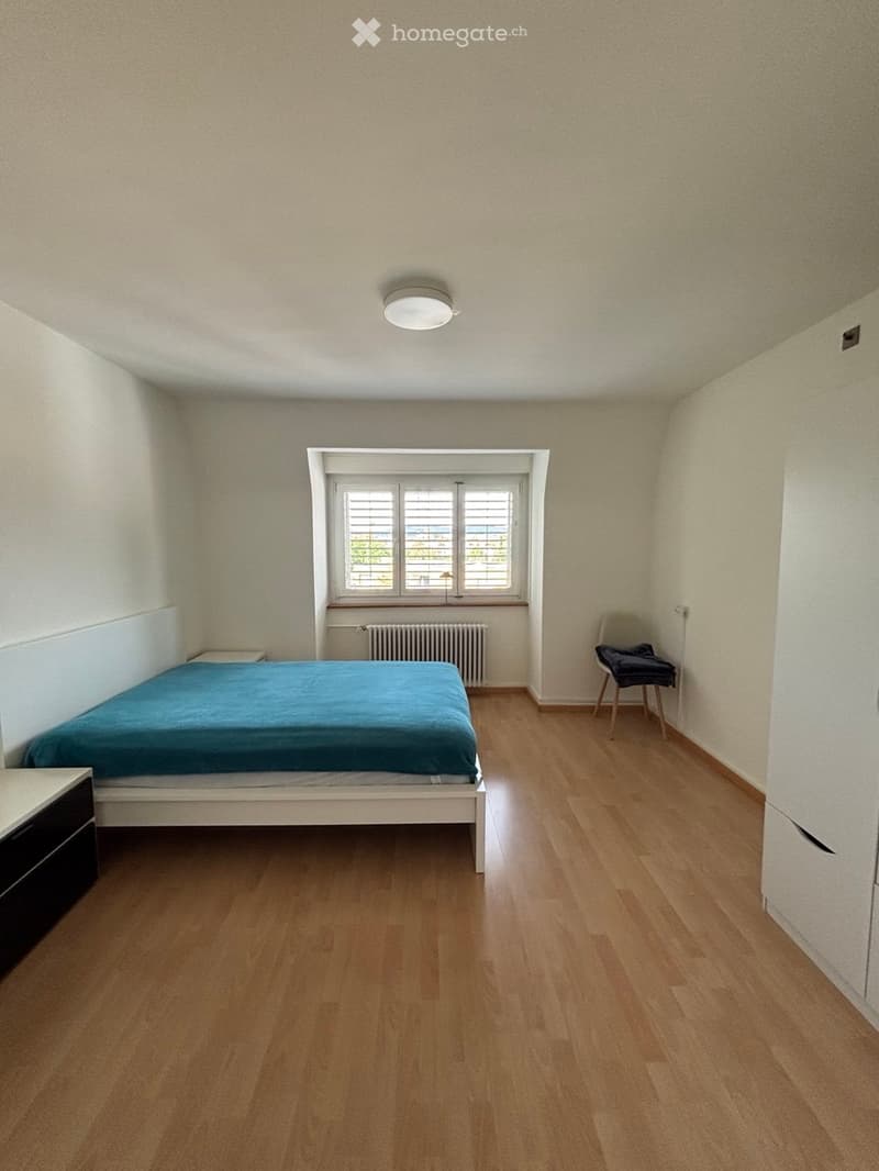 A furnished bedroom with a wooden floor, a single bed with blue bedding, a nightstand, a chair, and a radiator beneath a window.