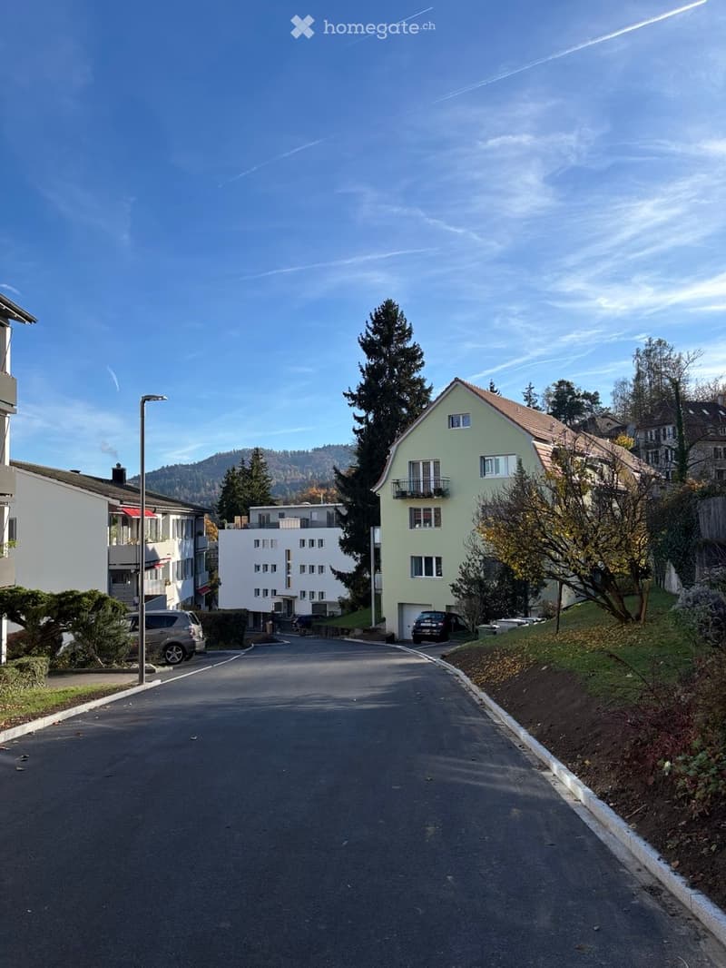 Residential street with multi-story houses, parked cars, trees, and a clear blue sky.