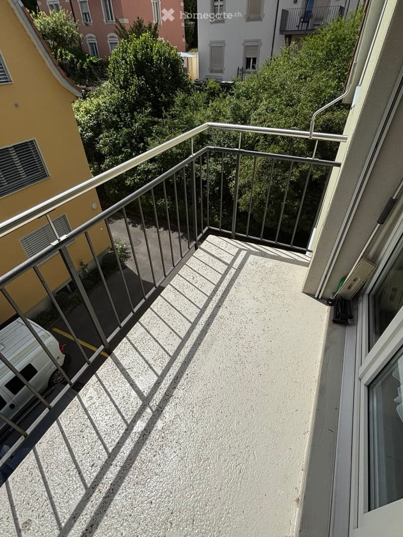 Balcony with metal railing, concrete floor, and view of neighboring buildings and greenery.