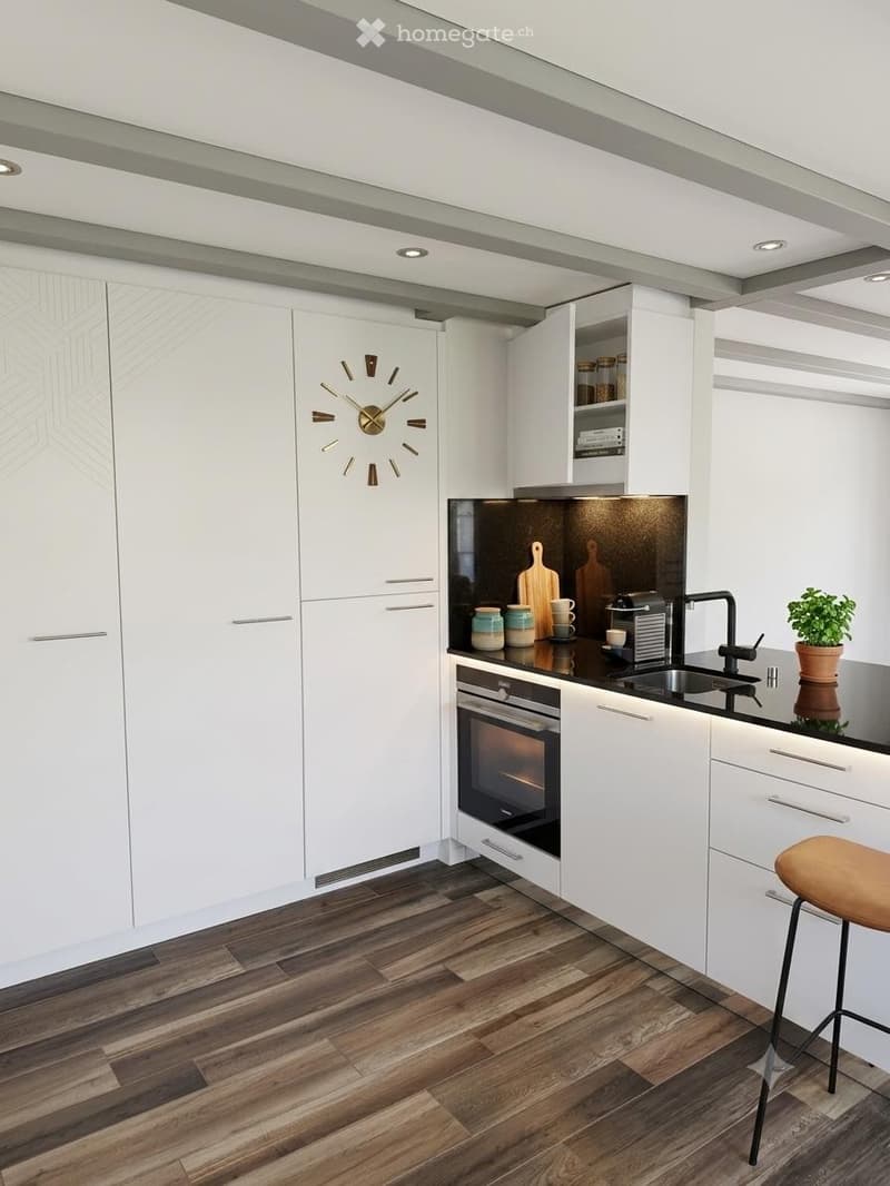 Modern kitchen with white cabinets, dark countertops, and wooden flooring. Features include an oven, sink, coffee maker, and a decorative clock on the wall.