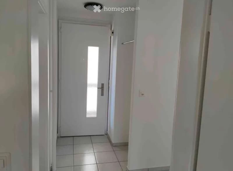 A narrow hallway with white tiled flooring, white walls, and a closed door at the end. The ceiling has a light fixture, and there is a small window on the door.