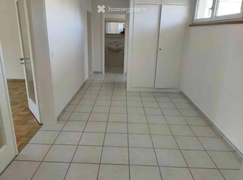 Empty hallway with tiled floor and white walls, leading to a small bathroom with a sink and mirror.