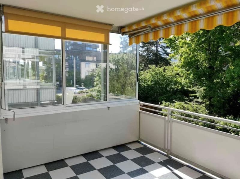 Balcony with checkered floor, large windows, yellow striped awnings, and a view of greenery and neighboring buildings.