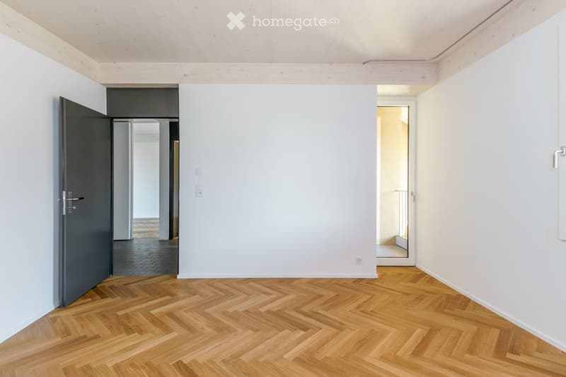 Empty hallway with light wooden herringbone flooring, white walls, and a dark door on the left leading to another room.