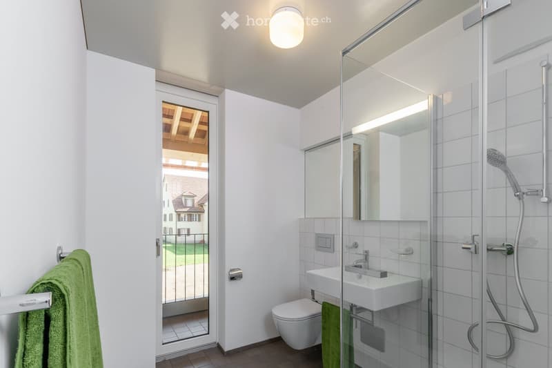 Modern bathroom with white tiles, a glass-enclosed shower, a toilet, a sink, and a towel on a rack. A large window offers a view of the garden and neighboring house.