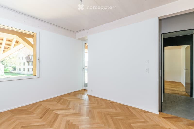 Empty room with white walls and herringbone wooden flooring, featuring a large window and a doorway leading to another room.