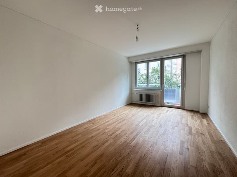 Empty room with wooden flooring, white walls, and a large window with a sliding door leading to a balcony.