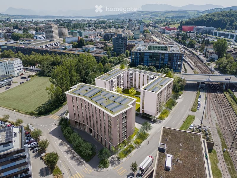 Aerial view of a modern residential complex with solar panels on the rooftops, surrounded by greenery, parking areas, and nearby urban infrastructure including roads, train tracks, and other buildings.