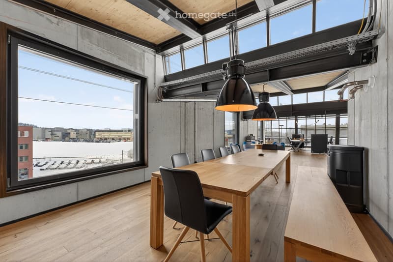 Modern, industrial-style room with large windows, wooden floor, long wooden table, black chairs, and hanging lamps.