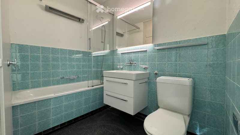 Modern bathroom with teal tiled walls, white fixtures including a bathtub, sink, and toilet, and a glass-enclosed shower area.