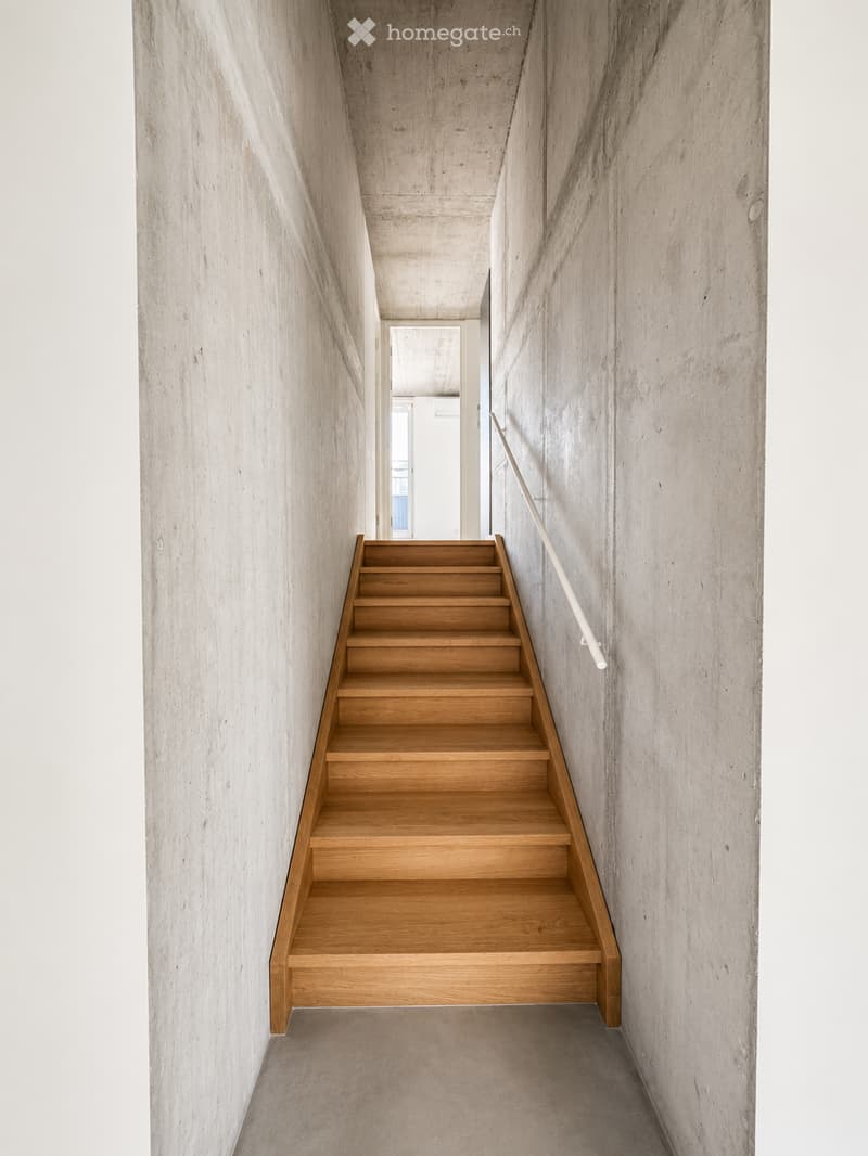 Modern concrete-walled corridor with a wooden staircase leading upwards, featuring a white handrail and a visible doorway at the end.