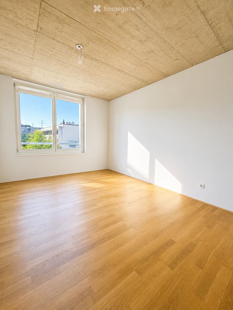 Empty room with light wooden flooring, white walls, and a large window letting in natural light. The ceiling is made of light wood panels.