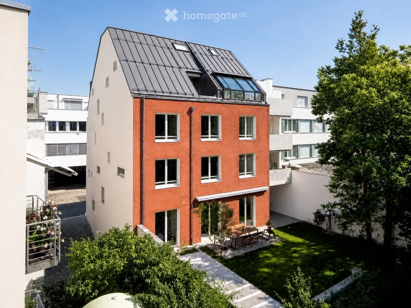 Modern two-story house with red-orange facade and grey roof, surrounded by greenery and neighboring buildings.