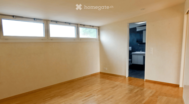 Empty room with wooden flooring and large windows, connected to a bathroom area.