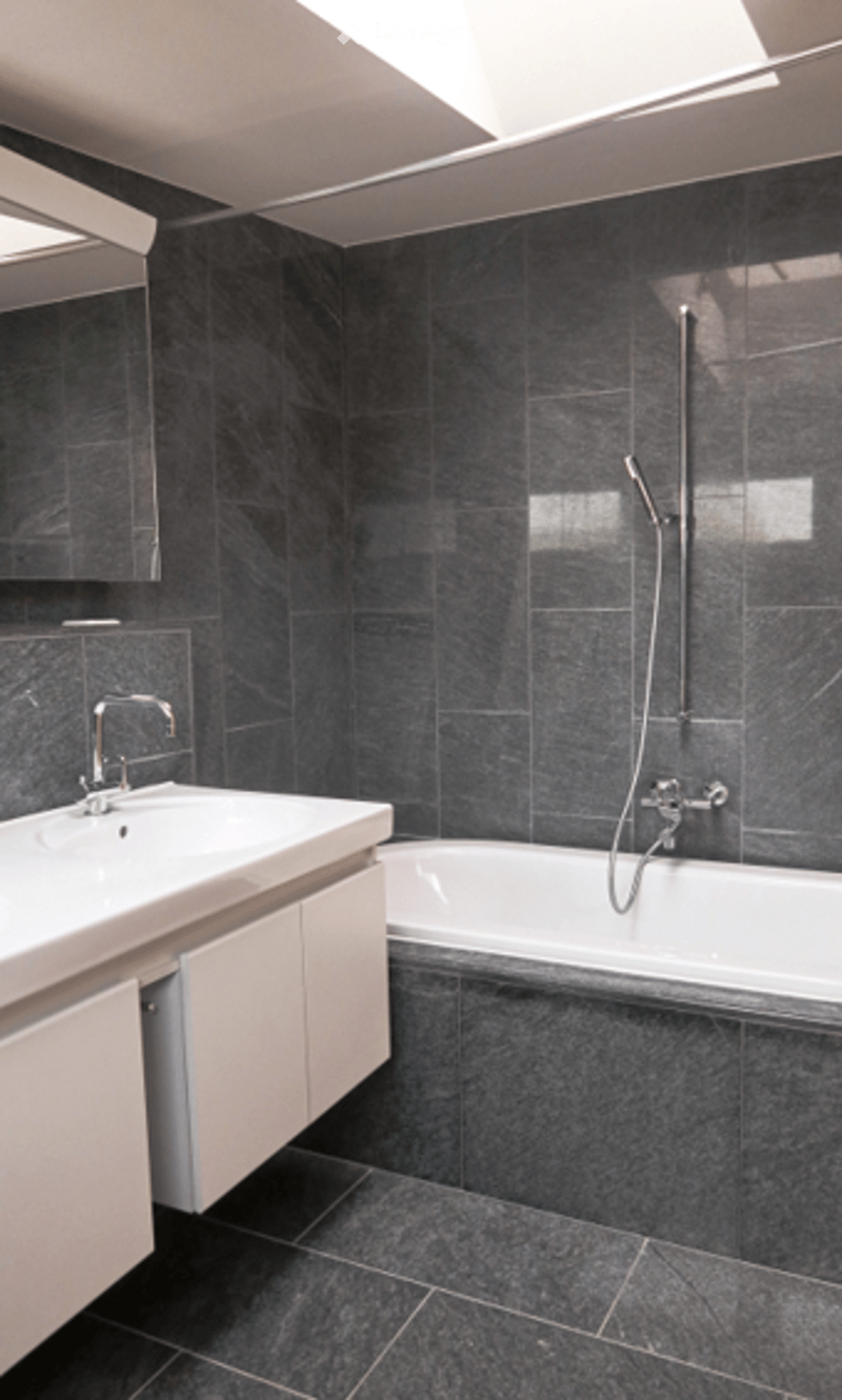 Modern bathroom with dark grey tiles, white sink, mirror, bathtub, and showerhead.