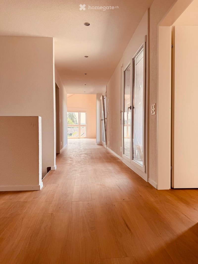 Bright hallway with wooden flooring, white walls, and recessed lighting. Features several doors and large windows allowing natural light to enter.