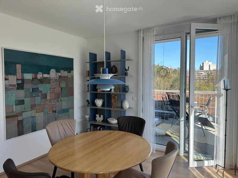 Modern dining area with a round wooden table, four chairs, a tall floor lamp, a large painting on the wall, and a built-in shelf unit with decorative items. Large glass doors open to a balcony with outdoor seating.
