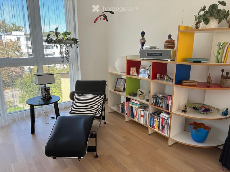 A modern, well-lit room with a wooden floor, a black lounge chair with a striped cushion, a small round table with a lamp, and a colorful wooden shelf filled with books, decorative items, and a radio. There is a large window with a view of greenery outsid