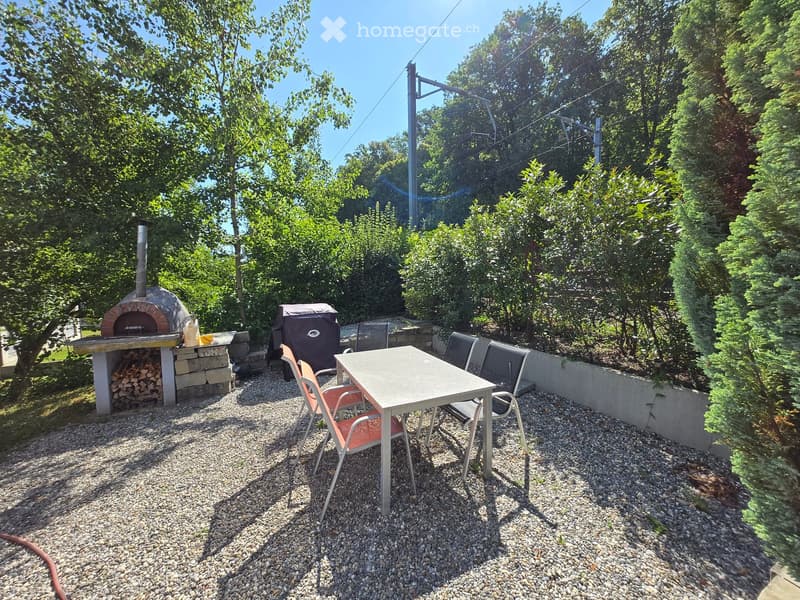 Outdoor patio area with a stone wood stove, a table with chairs, and a woodpile, surrounded by trees and greenery.