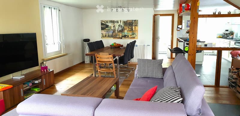 Bright living room with a grey sofa, coffee table, TV stand, dining table with chairs, and an open kitchen area with a counter, stools, and appliances.