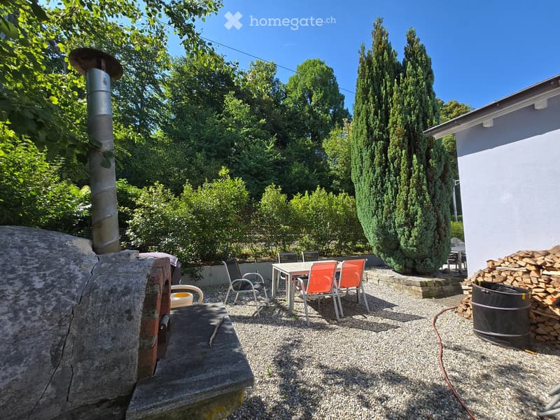 Outdoor patio area with a stone oven, a table with chairs, a woodpile, and a barrel, surrounded by trees and greenery.