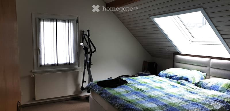 Cozy attic bedroom with slanted wooden ceiling, skylight window, bed with blue and green bedding, exercise bike, and a window with blinds.