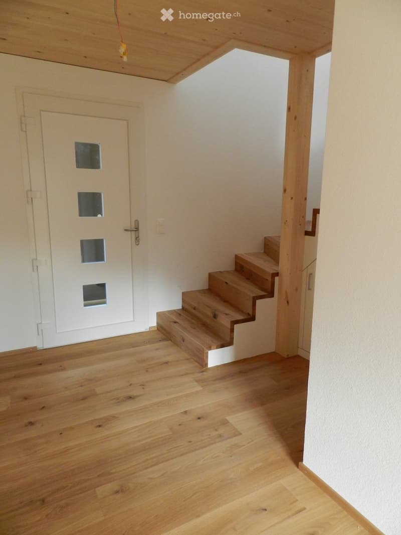 A narrow hallway with wooden flooring, a white door with glass panels, and a wooden staircase leading upwards.