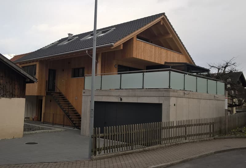 Modern two-story house with wooden facade, dark roof, garage, and balcony with glass railing.
