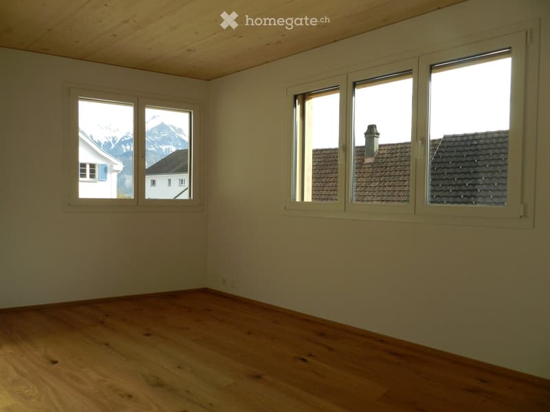 Empty room with wooden flooring, white walls, and large windows offering views of mountains and neighboring houses.