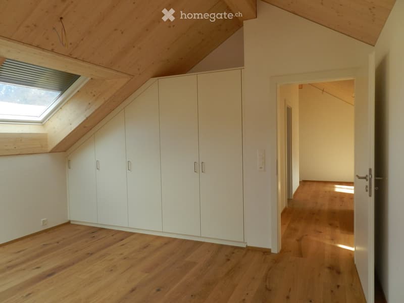 Bright, empty attic room with wooden floors, white built-in wardrobes, and a skylight window. The space features exposed wooden beams on the ceiling and a doorway leading to another room.