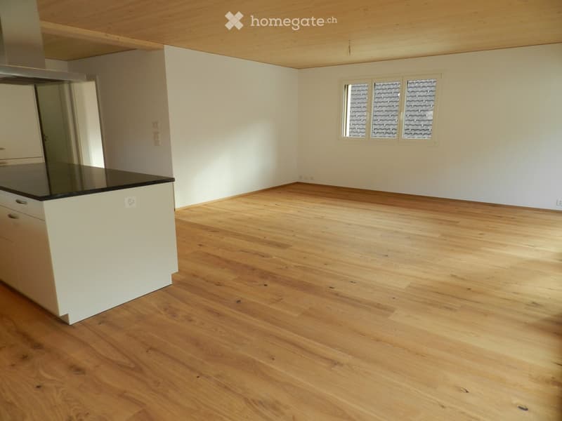 Empty kitchen with light wooden flooring, white walls, and a small island with a black countertop. Three windows with frosted glass panes are visible on one wall.
