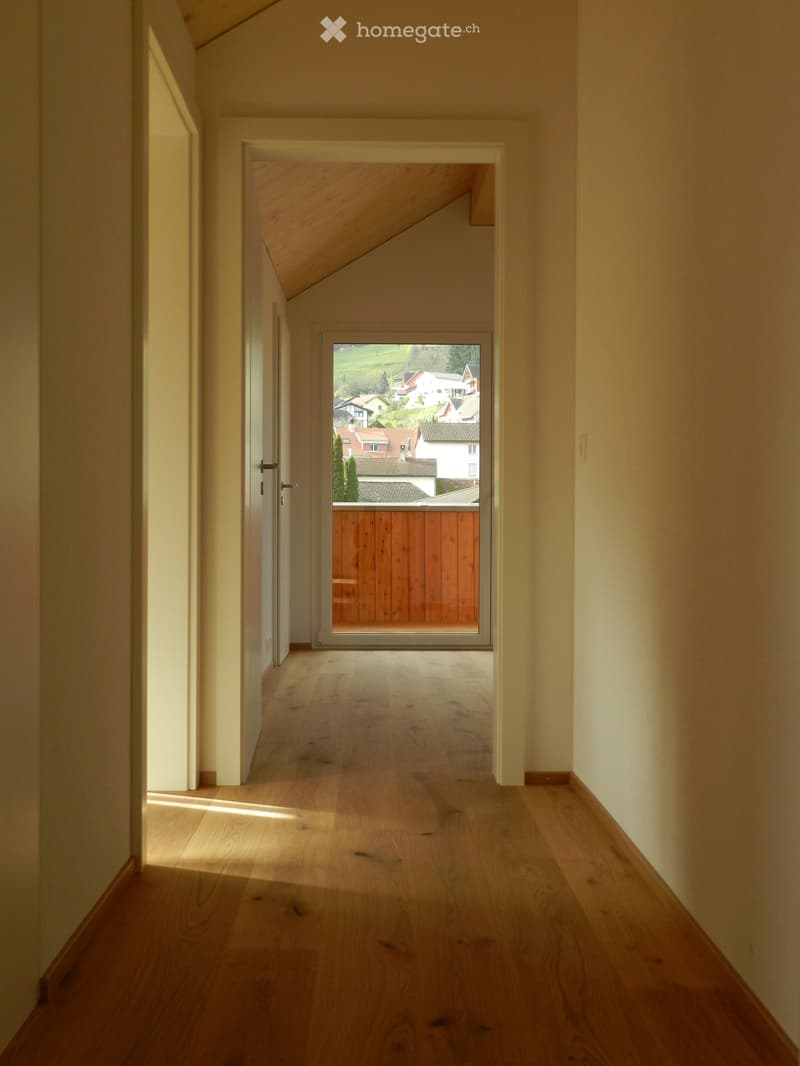A narrow hallway with wooden flooring and white walls, leading to a sliding glass door that opens to a view of a residential area.