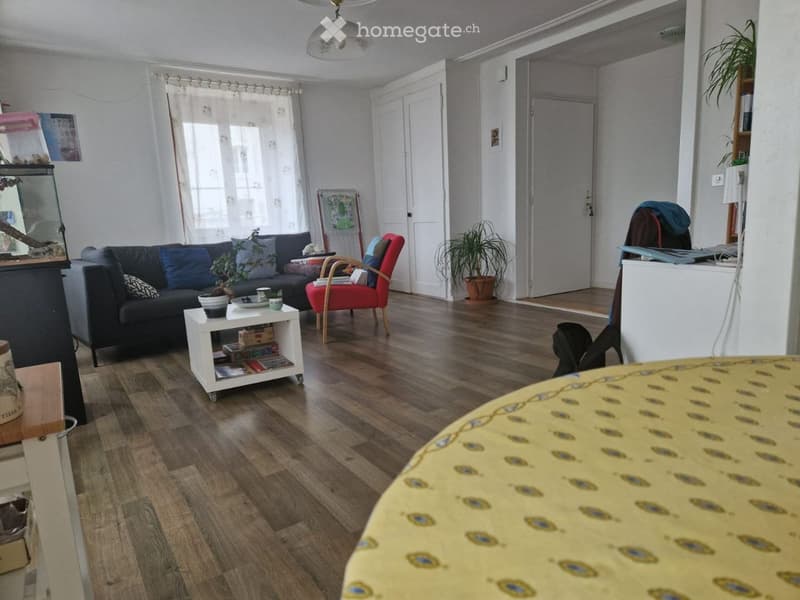 Living room with wooden flooring, a dark blue sofa, a red armchair, a small white side table, a fish tank, and a potted plant. The room has large windows with white curtains and a ceiling light fixture.