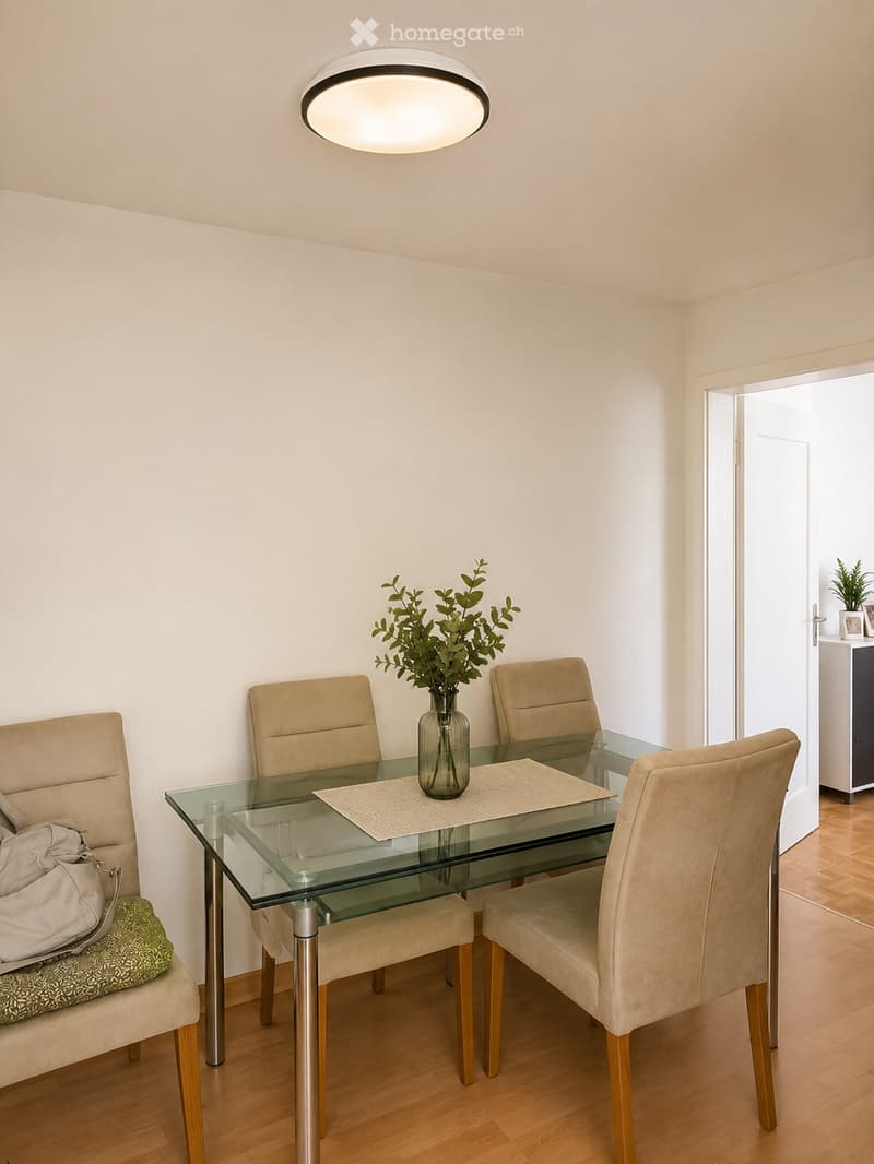Dining room with a glass table, four beige chairs, a vase with greenery, and a handbag on a chair.