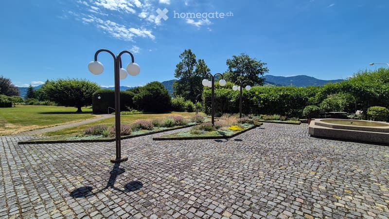 A well-maintained garden area with a cobblestone pathway, decorative street lamps, a small fountain, and neatly trimmed hedges and trees. The background features a grassy lawn and distant mountains.