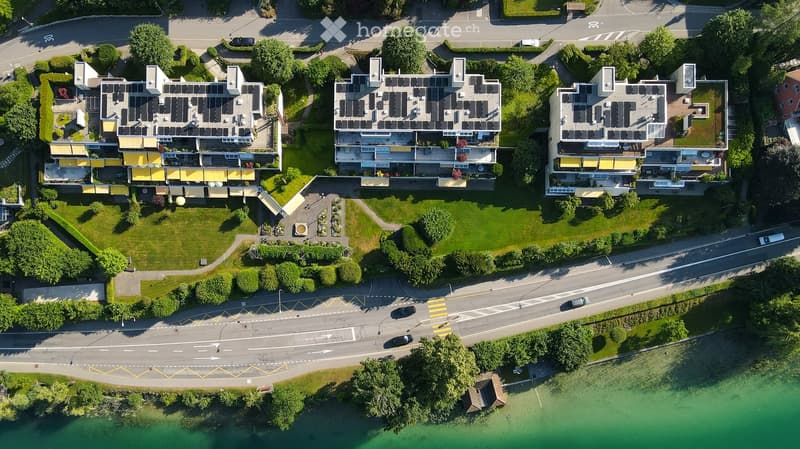 Aerial view of a residential complex with multiple buildings, solar panels on rooftops, and surrounding greenery. The complex is situated near a road and a body of water.